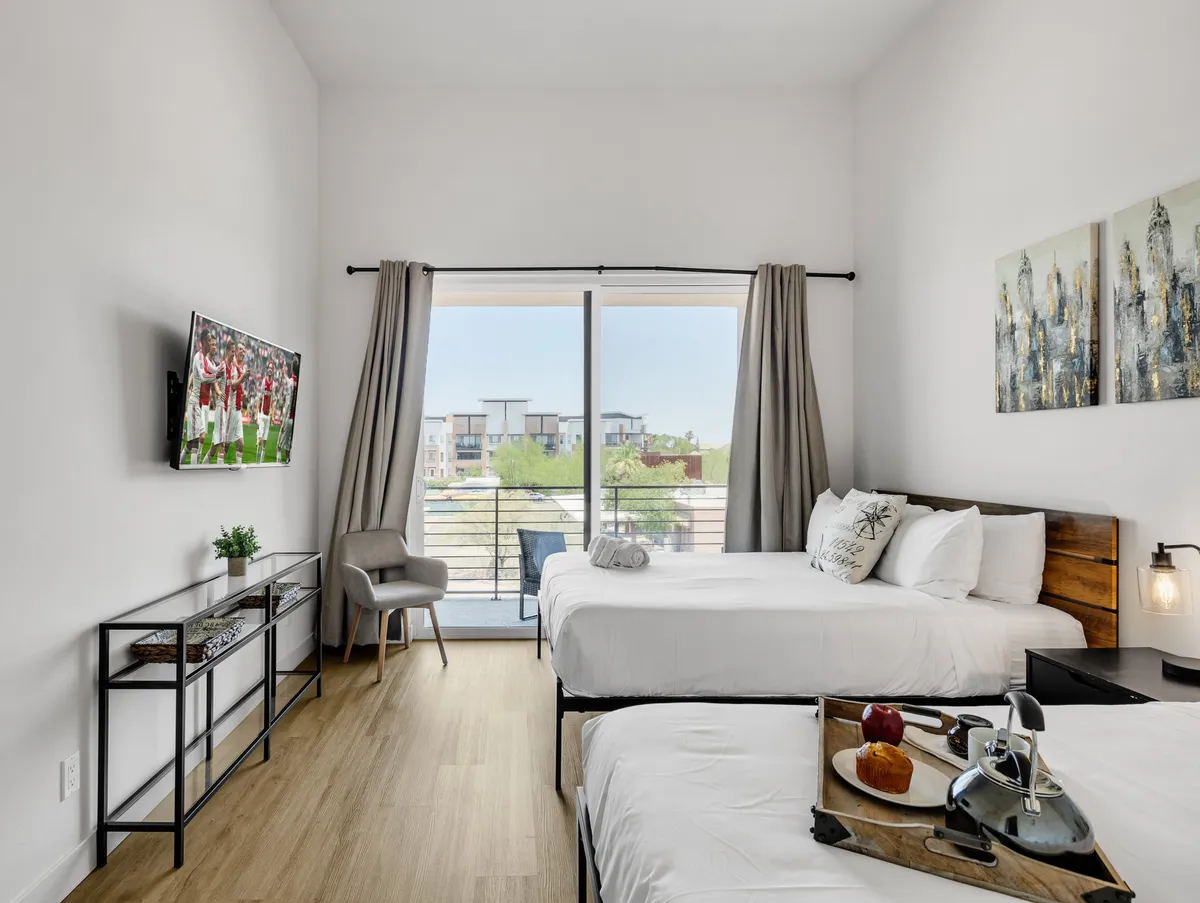 This image shows a spacious bedroom with two queen beds adorned with plush bedding. There's a large window leading to a balcony, with a TV mounted on the wall for entertainment. Decorative wall art and a small dining table with a tray complete the scene, suggesting both comfort and practicality for groups.