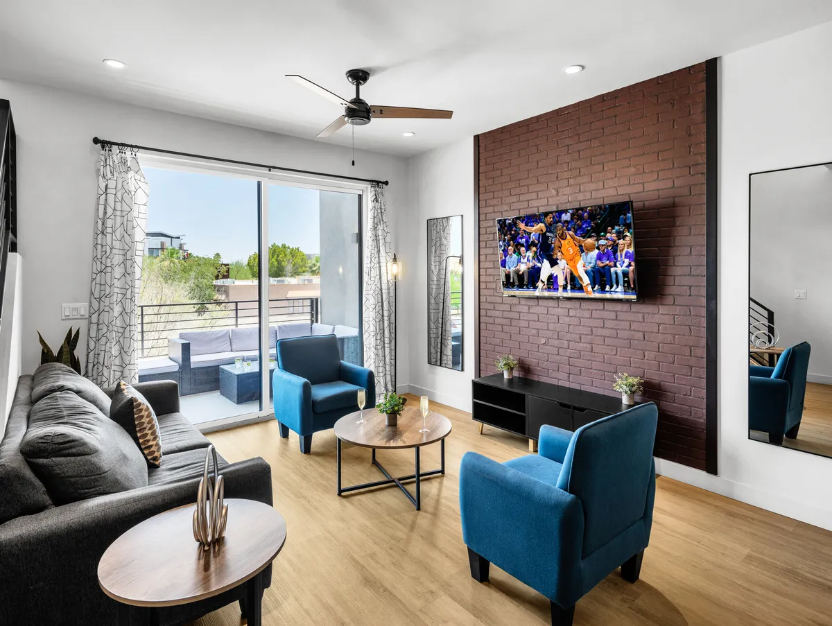 A cozy living area with a plush sofa, blue armchairs, a coffee table, and a smart TV dominates this scene. Large windows let in natural light, enhancing the sense of space and comfort.