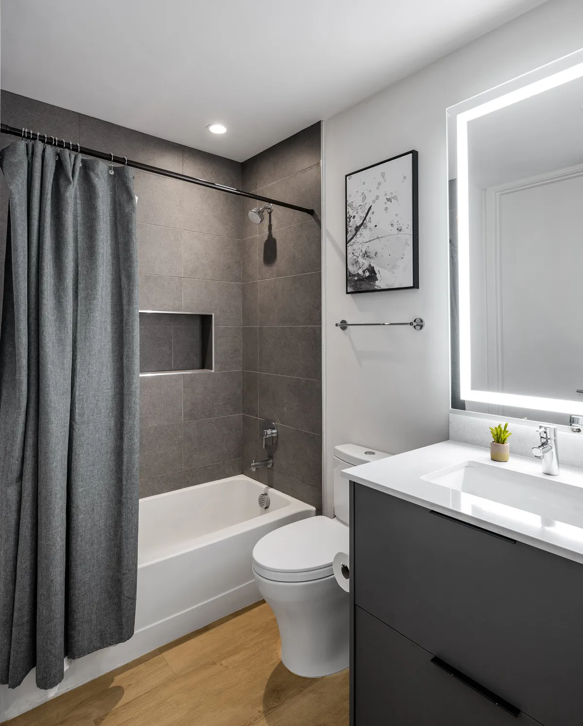 A modern en-suite bathroom equipped with a bathtub, a vanity with sink, framed wall art, and a shower curtain. The setup is bright, clean, and tranquil, ideal for relaxing after a long day or preparing for a night out.