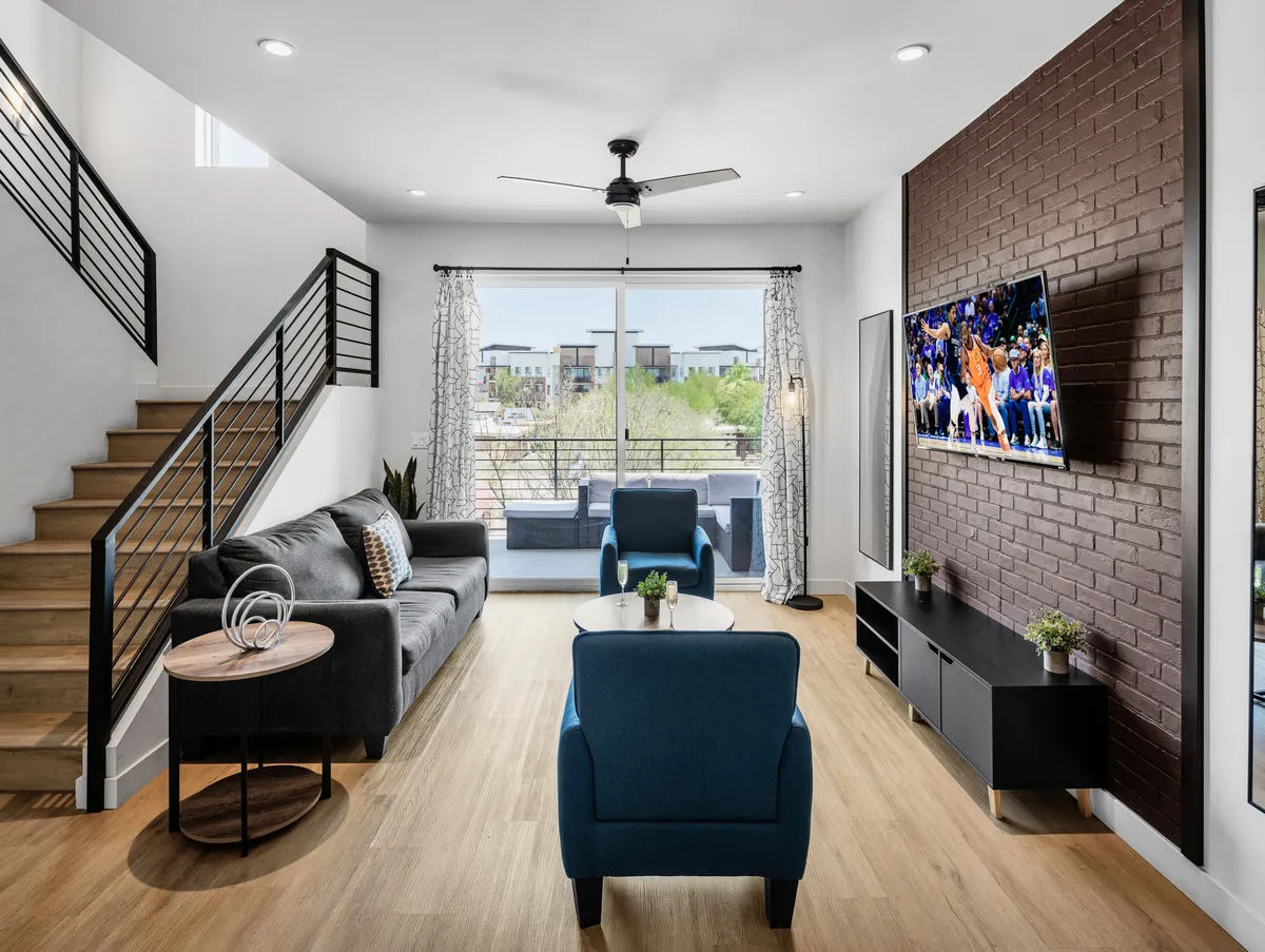 The image features a stylish, open-concept living room with a plush sofa, striking blue accent chairs, a flat-screen television mounted on an exposed brick wall, and a modern staircase leading up. The space feels inviting and perfect for gathering with friends or family, blending contemporary comfort with a welcoming, casual vibe.