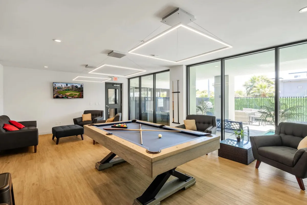 A stylish interior community clubhouse featuring a pool table, plush sofas, a large television, and abundant natural light from floor-to-ceiling windows. The space is arranged for social gaming, group entertainment, and relaxed gatherings.