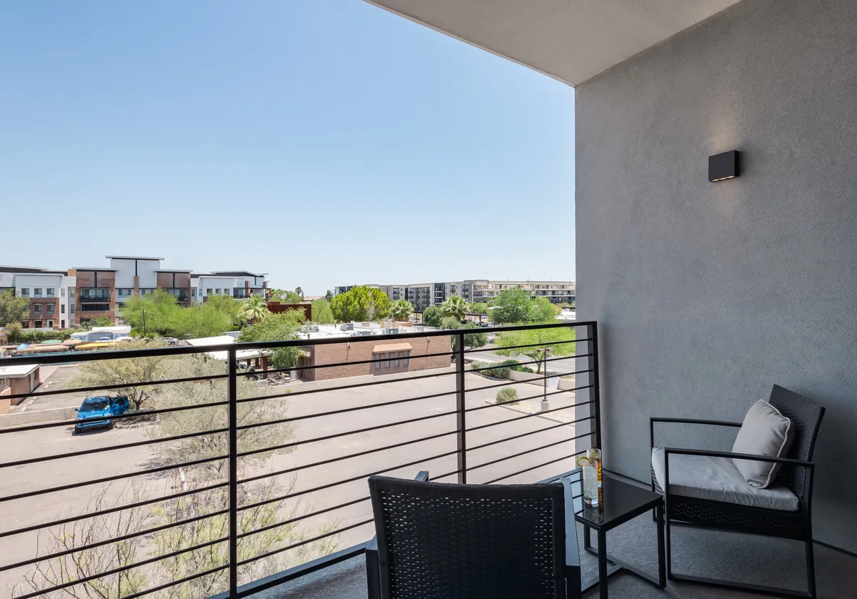 A private balcony furnished with seating overlooks the neighborhood, featuring calm railings and a relaxing ambiance. The view hints at both sunrise and sunset moments.