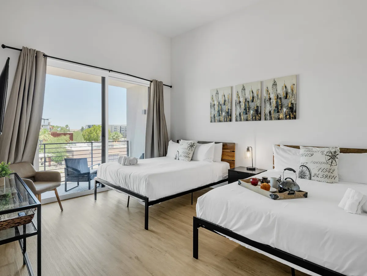 A serene bedroom with two inviting beds, decorative wall art, and a breakfast tray resting on one bed. The room opens to a balcony with outdoor views, indicating the presence of private outdoor space. Soft lighting and cozy bedding highlight the room's tranquil atmosphere.